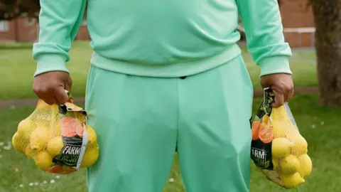 Archie Byron Man in green tracksuit holding lemons stood near grass. You cannot see his face or feet.