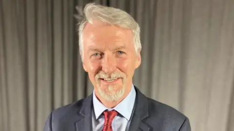 Huw Irranca-Davies, stood in front a curtain, wearing a dark blue suit, red tie, and blue shirt. He is smiling at the camera.