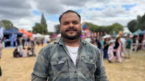 A close-up of a man in a checked green shirt with a silver chain, smiling at the camera. He has dark hair and a moustache. The background is blurred but a field with lots of stalls and crowds is visible.