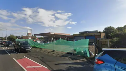 General view of a road with five cars driving towards to right of the frame. In the background there is a see-through green sheet supported by poles and behind it is a closed petrol station