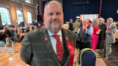 Labour leader Jim Robbins at an election count. He has a red Labour rosette pinned to a grey suit with light-coloured checks and a red tie. There are lots of people in a hall behind him.