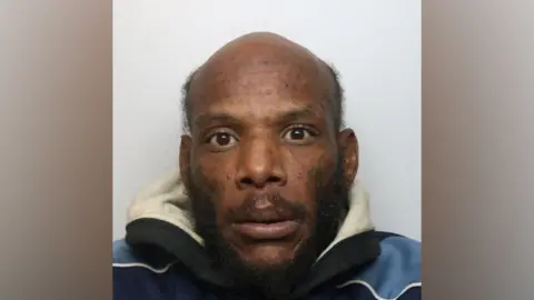 Tashaun Pink looks directly at the camera. He is partially bald, has cropped black hair on the side of his head and black facial hair. He is wearing navy and light blue jacket. 