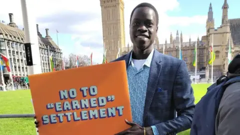 Omot Otor. He is standing outside the Houses of Parliament and is wearing a blue blazer and shirt. He is wearing an orange sign which reads, No to Earned Settlement.