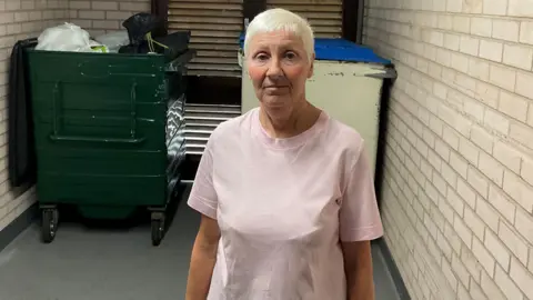 A woman with short white hair and wearing a pink t-shirt standing in front of two large bins. One has some white and black bags in and the other is closed. 