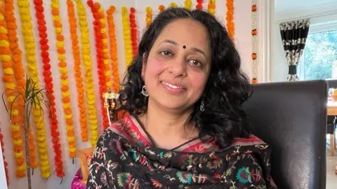 Rashmi Bansal wearing a black and multi-coloured shawl with orange, red and yellow pom pom garlands hanging on the wall behind her