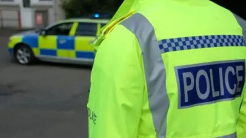 Stock image of a police officer pictured from behind, wearing a yellow high-viz police jacket. A police car can be seen, slightly out of focus, in front of them.