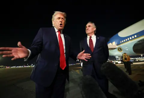 US President Donald Trump on the airport tarmac with Air Force One behind him. The president has his hands and mouth open as he speaks to a boom mic. Secretary of the Interior Doug Burgum stands to the president's left and watches him speak. 