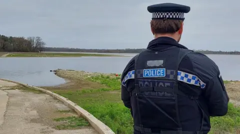 Essex Police A police officer with his back to the camera as he stands in front of a reservoir. He is in police uniform with a hat. It is a grey cloudy day. 