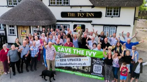A group of villagers standing outside a pub with a sign saying 'The Ivy Inn'. They are waving their hands in the air, some with thumbs up. There is a large green and white banner saying 'Save the Ivy'. The pub is painted white with a thatched roof