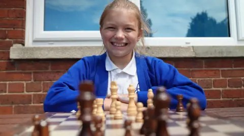 Evie has blonde hair and is smiling, in front of her is a blurred view of brown and white chess pieces at the board she is playing at. She is wearing a blue cardigan and a white shirt. 