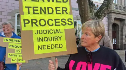 Sally Minty-Gravett holds a placard saying "Flawed tender process-Judicial inquiry needed"