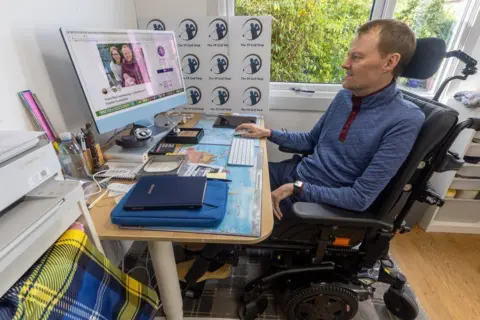 My Name'5 Dodgy Foundation Scott is working on the computer at his desk in a specially adapted chair. He is looking at a computer screen and is wearing a blue jumper.