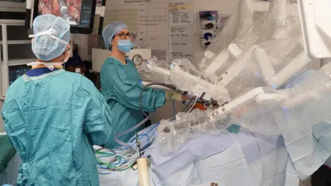Shaun Whitmore/BBC Two theatre staff watch over the multi-million pound robot which is being used to operate on Nick Samiotis. They are wearing turquoise theatre gowns and are adjusting one of the instruments that slots into the robot. The robots arms are like a white mechanical spider, reaching down into the patient. The two theatre staff are wearing masks and hair nets. The images from inside his body are projected onto a screen to their top left.