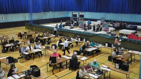 Dozens of people are counting votes in ballot boxes in a gym hall as part of the 2020 Election in Guernsey