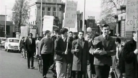 A black and white archived photo showing a group of protesters marching thorough the city centre holding placards demanding equality. One at the forefront reads 'love thy neighbour, let him work'. The people visible in the crowd are all men, dressed in blazers, suits and long jackets.