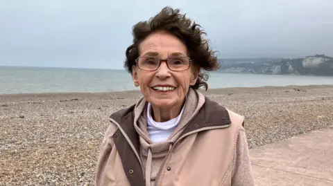 Joan Norcombe smiling while walking along Seaton seafront on a cloudy day. She is stood on an esplanade in front of a stony beach.