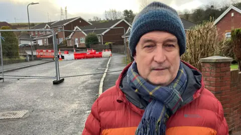 Simon Williams is standing outside the cordoned off road, wearing a red and orange winter coat, tartan scarf and blue and grey striped woolly hat. Metal barriers and a second orange and white safety barrier can be seen behind him.