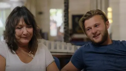 Jo and Sam sitting together in a hotel lobby during Race Across the World as Sam smiles and talks to fellow contestants off-camera