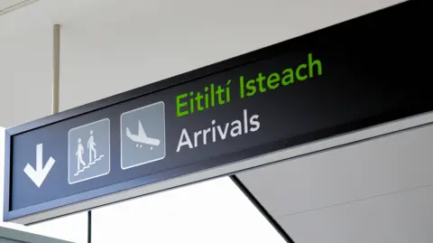 Getty Images A black sign reads "arrivals" with the Irish translation written above in green. Beside the text is an image of a plane, and people descending a staircase beside a white arrow pointing downwards