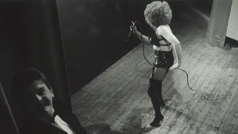 Ross Williams Black and white photograph form the 1980s with a drag artist on stage in a black leather outfit with a man dressed in a dinner suite in the wings looking at the camera