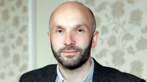 A man in his forties with a shaved head and a short beard. Flowery wallpaper is behind him and he is wearing a suit.