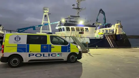 A police car is parked at Lerwick Harbour. A fishing trawler is moored nearby. Steps have been positioned leading from the harbour to the boat.