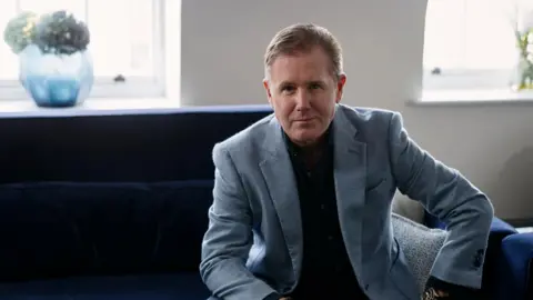 Rob Smalley/The Rigby Group A man with short dark blonde hair sits on a dark blue sofa wearing a light blue suit and a dark shirt.
