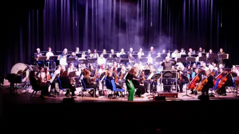 City of Wolverhampton Council A wide shot shows the full orchestra on a blue-lit stage with a soloist standing in the centre wearing green trousers. Stage smoke drifts across the performers while the silhouettes of hundreds of audience members are visible in the foreground. 