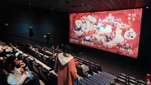 Getty Images Back view of people at a theatre. On the screen is a poster for Ne Zha 2