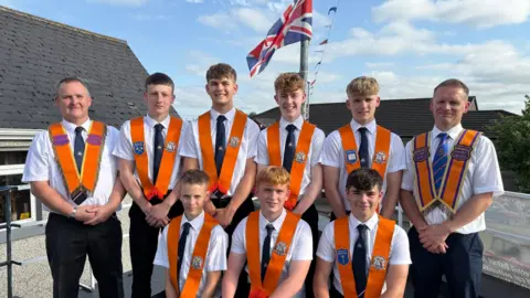 Seven teenage boys standing smiling at the camera - either side are two male leaders. All the men are wearing orange sash's, navy ties and dark suit trousers. There is a blue sky with a Union Flag flying in the middle.