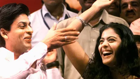 Reuters Actor Shah Rukh Khan and actress Kajol smiling and cheering, with Shah Rukh in a white shirt tapping Kajol's raised elbow