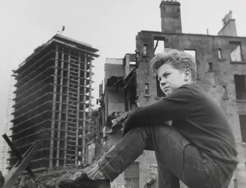 Getty Images A black and white image of a young Gorbals boy sitting on a wall in front of a tenement in the middle of being demolished, with the bones of a tower block under construction beyond it.
