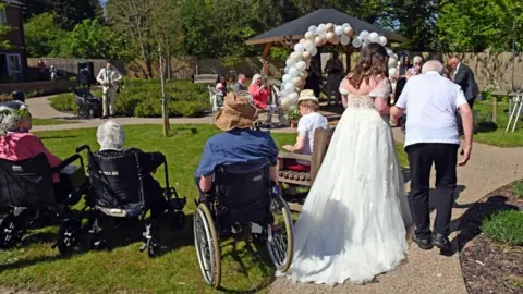 Emma Noake Emma in her wedding dress is accompanied by George in a white top as they walk down a path. On the left, people in wheelchairs can be seen looking at a gazebo with a balloon arch formed at the entrance.