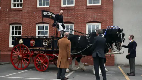 Getty Images A horse with a fancy cart behind it and surrounded by well dressed men. The Wadworth logo is painted on a black arch over the top of the cart