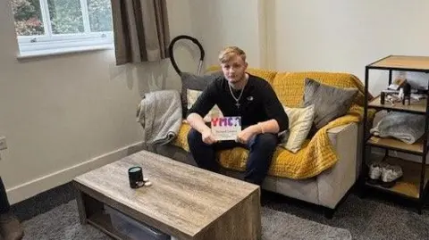 A young man with blonde hair and black clothes sat on a small sofa in front of a low table in a grey-carpeted room