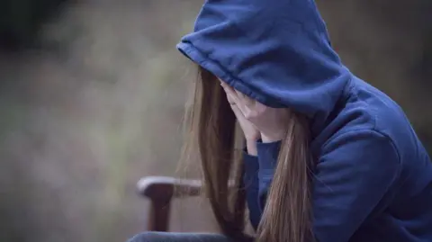 A young girl sat on a bench with her head in her hands. Her dark hair is hanging down the side of her hands. She is wearing a blue hoodie with the hood over her head. The backdrop, which appears to be outside, is out of focus.