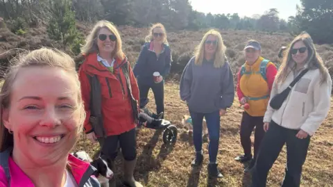 Katrina Emerton Katrina Emerton takes a selfie and smiles at the camera. She has brown hair that is tied up and wears a pink jacket. Behind her are five other women who all also smile at the camera. They all wear outdoor clothing and one stands next to a dog on a lead while another holds on to a pushchair.