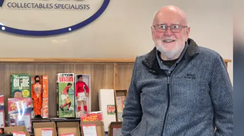 Vectis Auctions Bob Brechin, a bald man with a white beard wearing a grey zipped jacket, stands in front of a display of toys including Action Man and GI Joe figures
