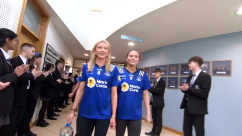Two girls wearing blue sports kits walk along a school hallway. One is holding a trophy in her right hand. School pupils wearing black uniforms flank them on either side. The pupils are clapping.