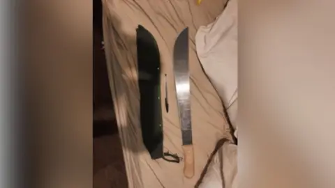 West Yorkshire Police A machete knife lying next its sheath on a blanket of some description. A biro pen sits between the knife and the sheath.