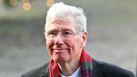 PA Media Kenny MacAskill smiles off to the side. He has short white hair and wears silver glasses. He wears a dark coat and white shirt and has a tartan scarf around his neck.