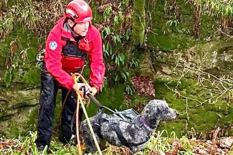 LMRT Member of LMRT in red jacket and red helmet, attached to climbing ropes with a small black curley haired dog on a harness, standing on a grass covered bank