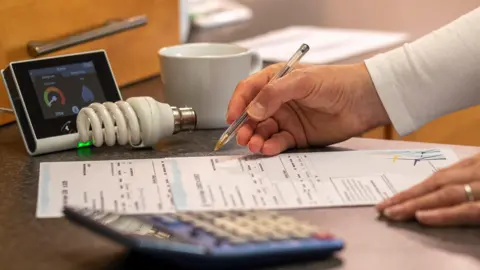 Getty Images A person looks at their bills at home. A pen is in one hand and a bill lies on a table. There are other objects on the table, like a calculator, a meter, a bulb and a cup