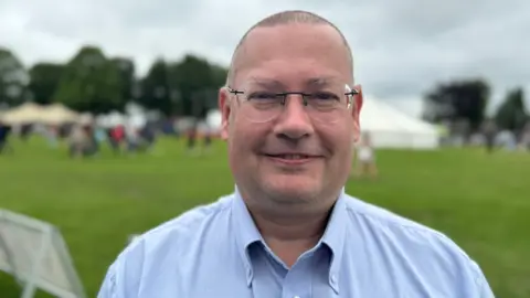 Simon Outen-Coe pictured standing on a grassy area. He is wearing spectacles and has a blue shirt and very short hair. He is smiling at the camera.