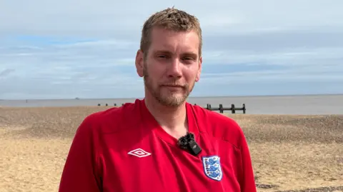 Luke Deal/BBC A head and shoulders image of a man with a red England shirt on. He is standing on the beach with the sea behind him.
