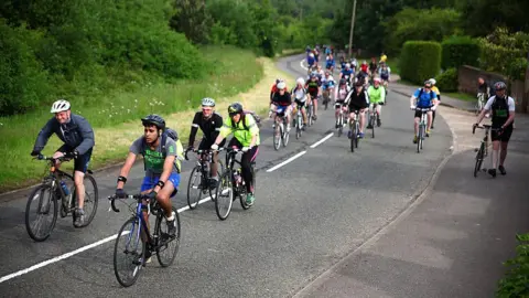Dozens of cyclists pedal up a hilly road. A couple have dismounted and are walking on the pavement. The road is surrounded by greenery.