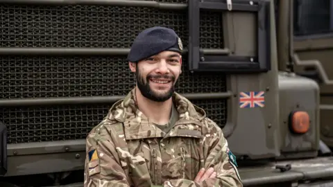 L/Cpl Lee Moulton wearing an Army jacket with a camouflage pattern in browns, cream and muddy green. He is also wearing a black beret with an insignia just seen on his right side. He has a short black beard and is standing with his arms folded and smiling, while behind him can be seen the front grille of an Army truck.