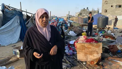 A Palestinian woman looks down towards the camera as she inspects damage to a camp housing displaced people in Khan Younis, after an Israeli strike on 2 July. Other people, broken tents and strewn belongings can be seen in the background against a blue sky.