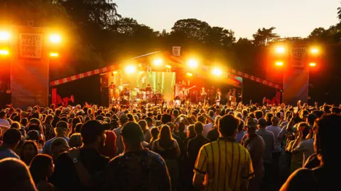 Crowds gather at a stage with artists playing. Signs on either side of the stage say "Mostly Jazz, Funk and Soul Festival". Orange lights are flashing as the crowd plays