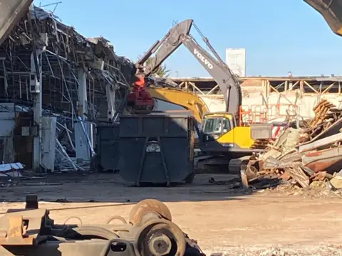 Luke Deal/BBC A digger is working on demolishing the former Delphi building. Parts of the building have been torn down with twisted metal visible and debris on the floor